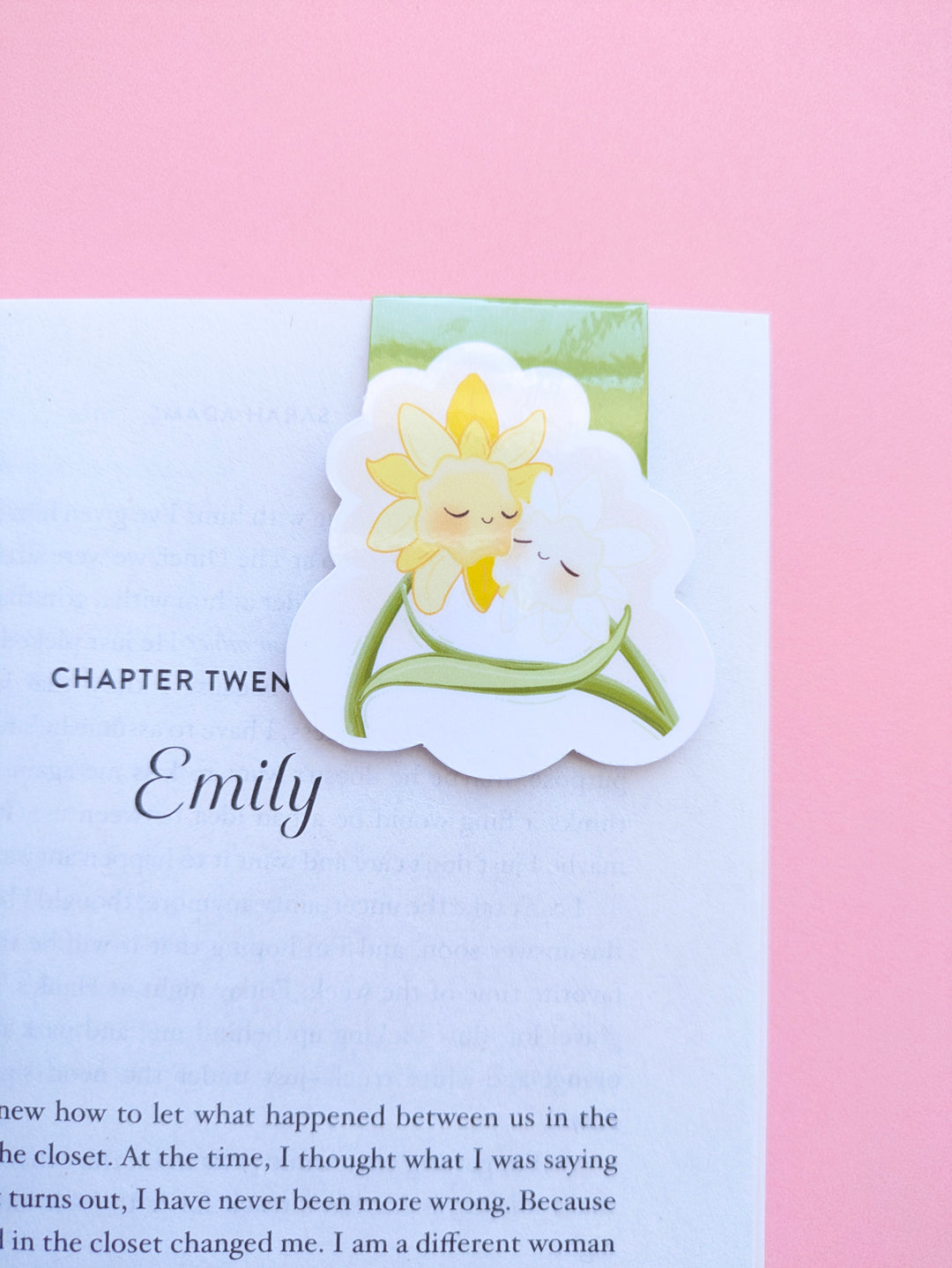A zoomed in, high aesthetic reading setup featuring the Daffodil Hug Szn 'Meadow Green' magnetic bookmark clipped to Sarah Adams’ 'Beg, Borrow, or Steal.' The sage green kawaii character bookmark is positioned on a crisp white page against a rose pink background. Light reflects off the high-density gloss finish, highlighting the secure, no-slide grip. Perfect for book annotating and curated reading rituals.