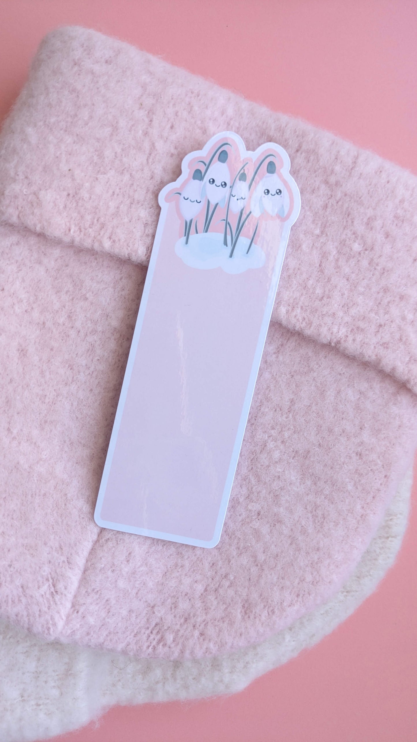 A Kawaii Sleepy Snowdrop 'Icy Venusian Pink' die-cut bookmark centered on an elegant soft pink wooly winter hat. The shot captures multiple reflective gloss sheen points across the laminated surface, highlighting the Snowdrop White characters and sage-silver leaves. A hint of an off-white scarf underneath completes this quintessentially kawaii, cozy, romanticized winter reading accessory scene.