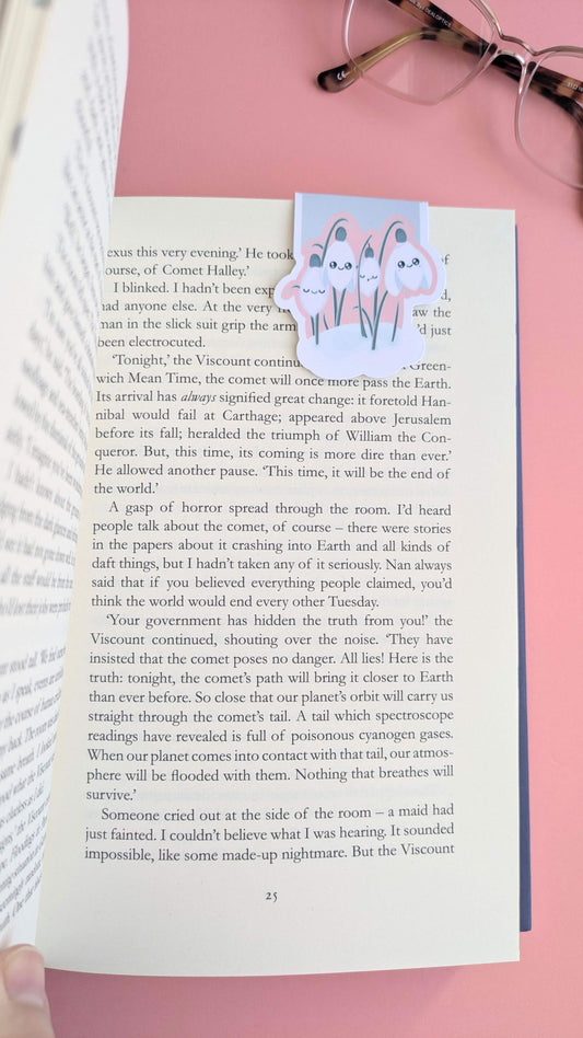 An aerial shot of an Ateu Studio Kawaii Sleepy Snowdrop 'Icy Venusian' pink magnetic bookmark on the right-hand page of a freshly opened book. A thumb is visible at the spine, capturing the moment of opening a new story, with the pages showing a natural lift at the bottom. The scene is styled with reading glasses on a rose-pink background in natural light, emphasizing the soft pink tones and the tactile, intentional experience of a new reading journey.