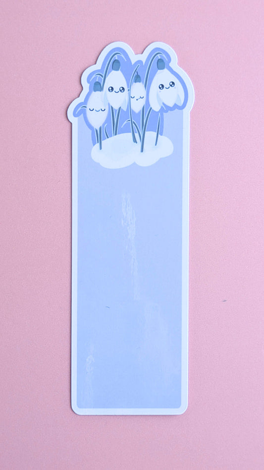 A direct aerial product shot of the Kawaii Sleepy Snowdrop 'Midnight Mist' lavender blue bookmark on a rose-pink background. The shot captures the premium gloss sheen of the bookmark as the light reflects off the durable laminated surface, highlighting the high-quality finish and the vibrant lavender-blue tones and Snowdrop White contracts of the character art.