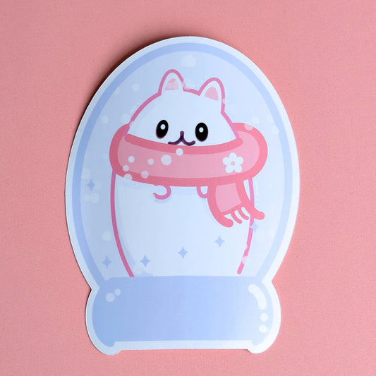 Kawaii Lil Beta character in a lavender snowglobe sticker with a chunky pink daisy scarf. Features swirling purple stars and translucent fluffy snowflakes. Designed by Ateu Studio for winter journaling and Kindles. Lil Beta, a minimalist egg-shaped cat sticker. The character features small ears, oval eyes, and a rounded 'w' shaped mouth, tucked inside a lavender winter snowglobe. Detail shows a chunky pink daisy scarf and pink paws and ears. High-quality gloss vinyl with signature highlight accents.