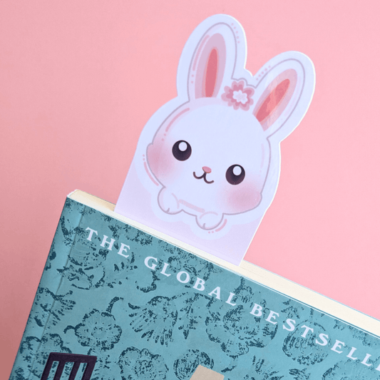 A close-up, angled view of the Marshmallow Blush Bunny Pink bookmark tucked into a closed copy of Before the Coffee Gets Cold. The vibrant pink and white bunny ears peek out from the spine, contrasting beautifully with the turquoise book cover and Ateu Studio pink background, highlighting the high-gloss finish.