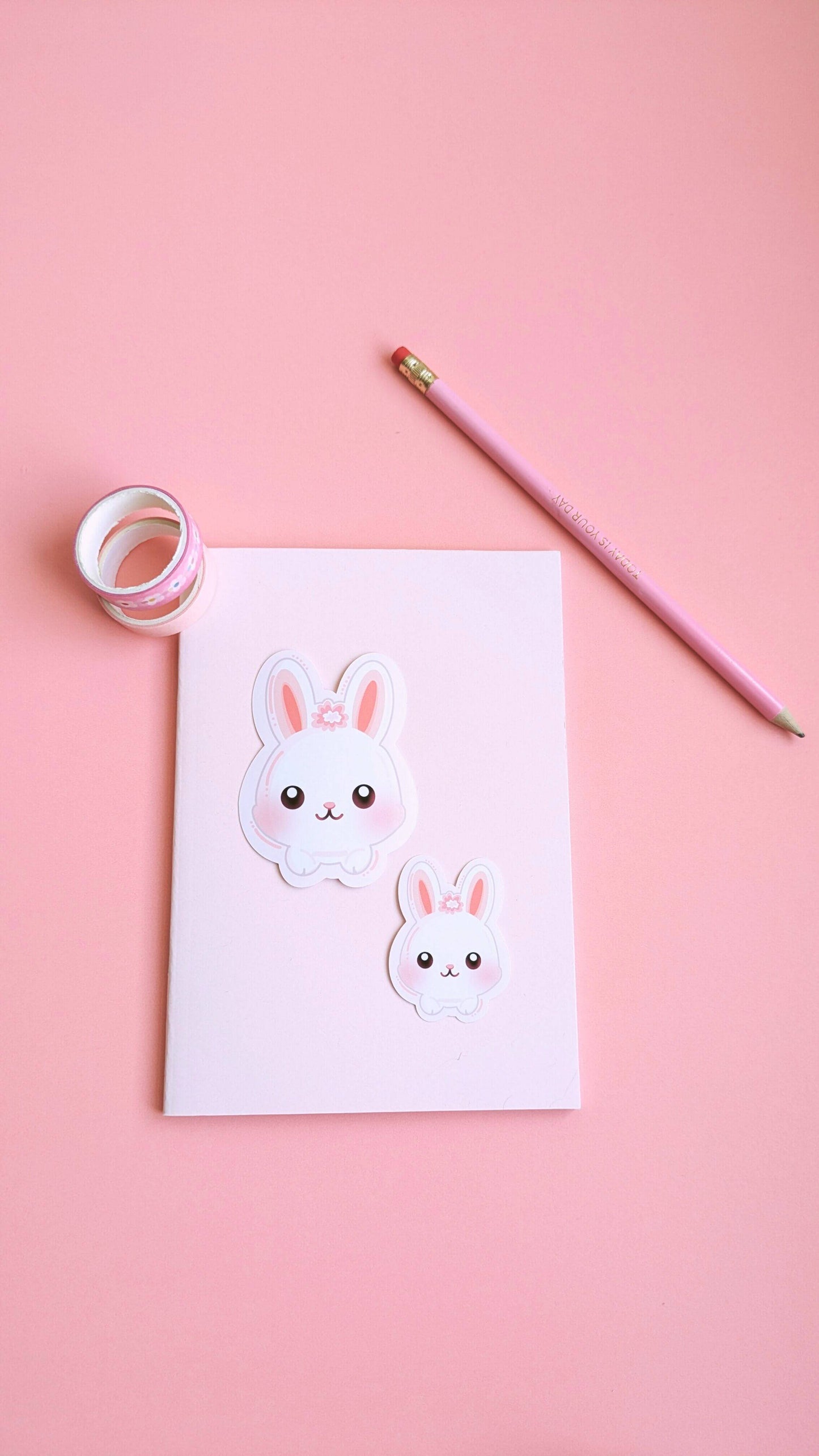 A vertical, minimalist view of a pale pink pocket notepad featuring two Marshmallow Blush Bunny stickers and a matching pink pencil. This linear composition highlights the 'Obsessed with Color' brand essence and intentional desk organization.