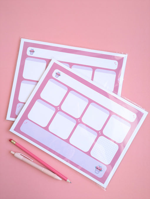 An arial shot of two 'Lil Buddy' weekly desk notepad planner that are in protective sleeves and laid over each other. there is a dark pink pencil and a pink on at the bottom of the image for scale and the background is a deep blush like pink. 