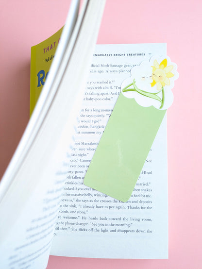 A wide aerial view of the Meadow Mist Green Daffodil Hug Szn kawaii bookmark inside an open book on a pink background. The die-cut top extends above the page, with natural shadows and a yellow book cover corner visible as the pages are opened for a reading session.