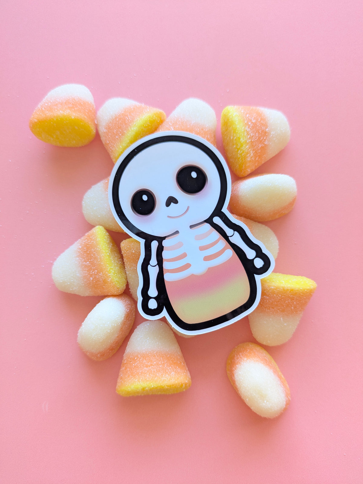 Skelicorn Dark gloss vinyl sticker, a kawaii skeleton with a white skull and bones over a pastel candy corn body, black outer detail and black eyes, surrounded by large fluffy marshmallow candy corn on a pink background.
