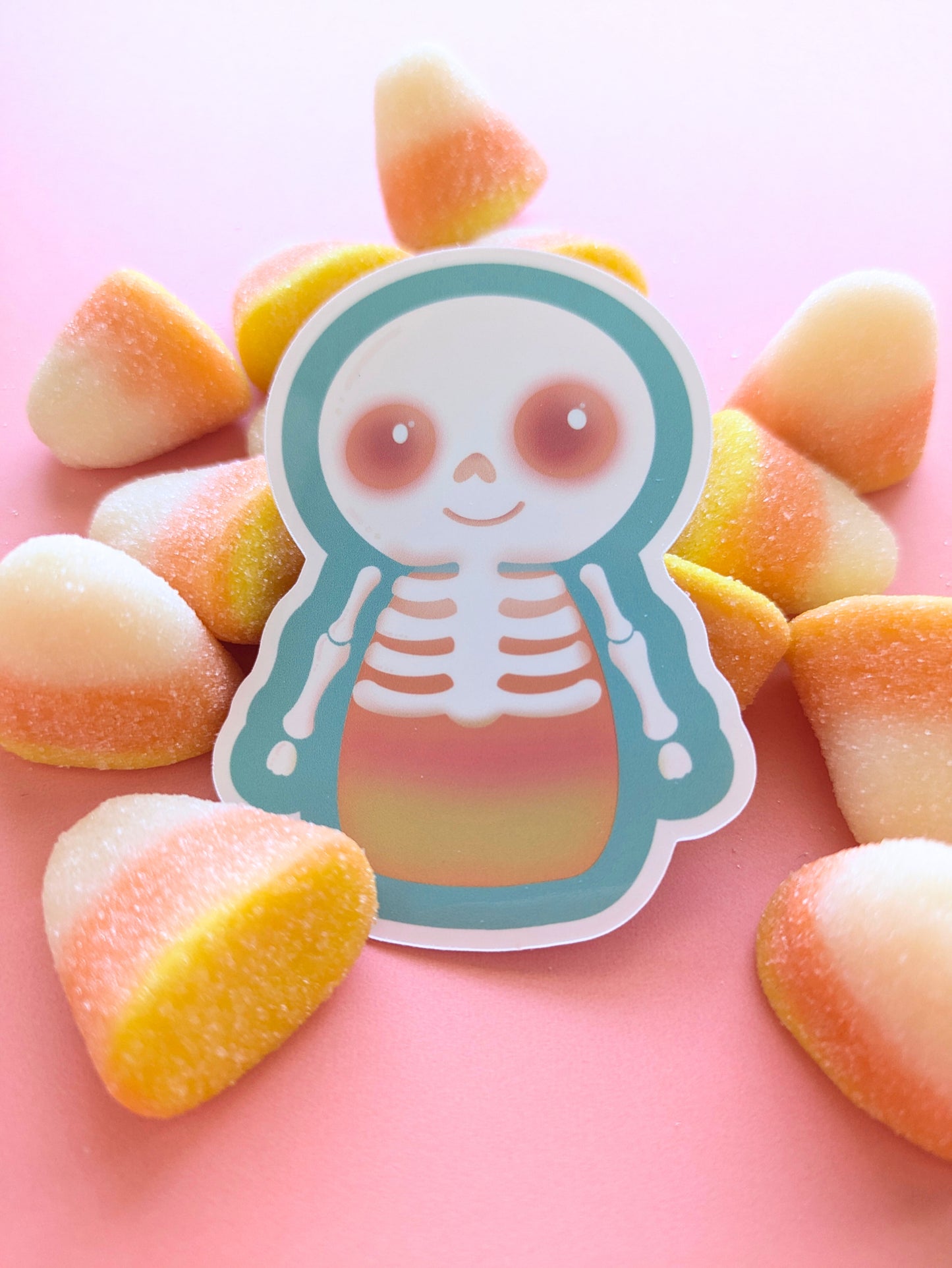 Skelicorn Luminous gloss vinyl sticker, a kawaii skeleton with a white skull and bones over a pastel candy corn body, turquoise outer detail and glowing Candy Coral pink eyes, surrounded by large fluffy marshmallow candy corn on a pink background.