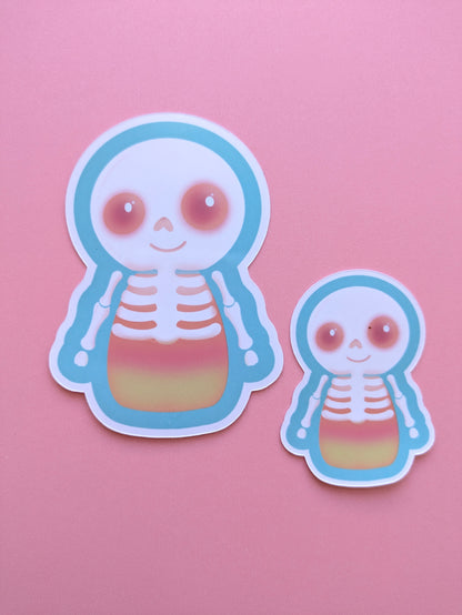 Two Skelicorn Luminous gloss vinyl stickers in 2 inch and 3 inch sizes, each featuring a kawaii skeleton with a white skull and bones over a pastel candy corn body, turquoise outer detail and glowing Candy Coral pink eyes on a pink background.