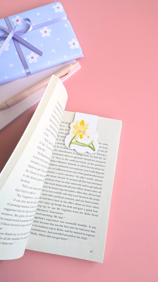 An aerial, naturally lit kawaii stationery composition featuring an open copy of 'A Quiet Place' by Seichō Matsumoto.  A Daffodil Hug Szn 'Daffodil White' magnetic bookmark is centered on the right page, casting a gentle shadow. Dominating the left third is a coordinated gift wrapped in soft blue daisy print paper with a satin ribbon, sitting near a pink mini notepad and pen. This color matched setup showcases a high-aesthetic, ready to gift reading ritual.