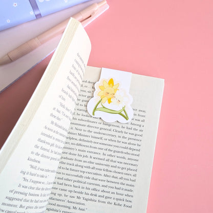 A detailed close up of the Daffodil Hug Szn 'Daffodil White' magnetic bookmark centered high on a curved book page. Natural light highlights the gloss finish and secure, no slide grip of the kawaii characters. A pink notepad, pen, and a hint of the blue daisy gift wrap are visible in the soft focus background, emphasizing the premium material quality and intentional color matching of Ateu Studio.