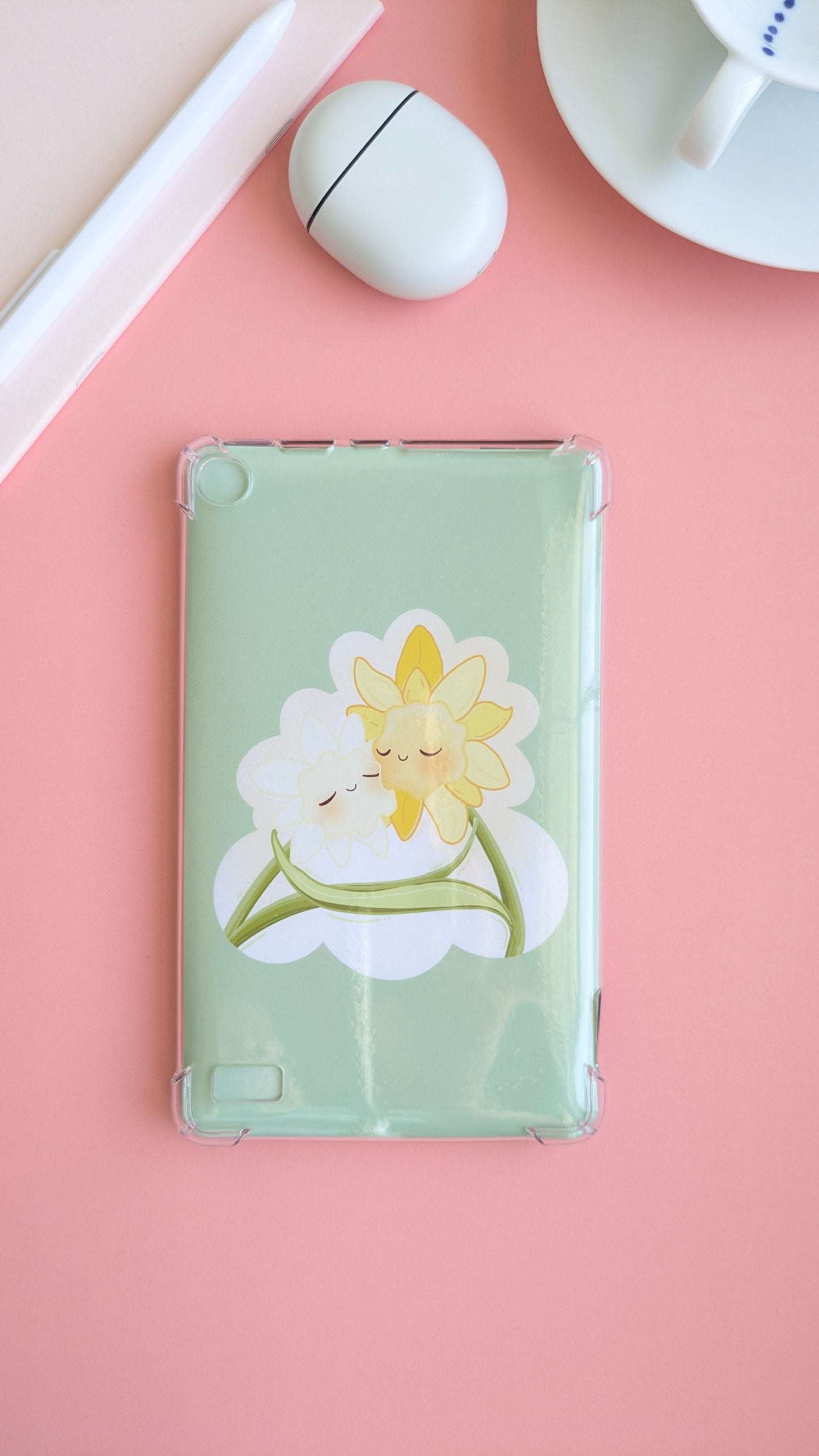 An aerial, naturally lit Kindle setup featuring a 'Soft Glow' green kawaii Daffodil Hug Szn insert. The design mirrors an oversized die-cut sticker, creating a high-aesthetic 'tech-scape' on a pink desk. Surrounded by a light pink notepad, white pen, Pixel Buds case, and a fine porcelain saucer, the high density gloss finish reflects light for a premium, color matched look.