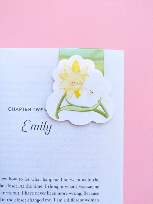 A zoomed in, high aesthetic reading setup featuring the Daffodil Hug Szn 'Meadow Green' magnetic bookmark clipped to Sarah Adams’ 'Beg, Borrow, or Steal.' The sage green kawaii character bookmark is positioned on a crisp white page against a rose pink background. Light reflects off the high-density gloss finish, highlighting the secure, no-slide grip. Perfect for book annotating and curated reading rituals.
