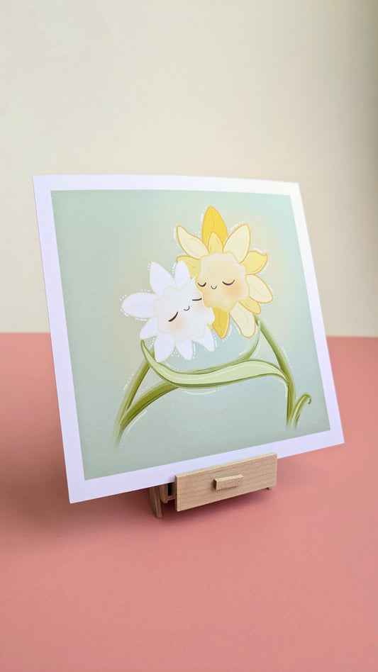 A 5x5 'Daffodil Hug Szn Meadow Mist Green Mini Art Print' on a white desk at an angle, with a white wall behind it. The original artwork by Ateu Studio features kawaii daffodils greeting each other with a hug after a long winter against a warm yellow-green spring sky. The print is resting on a miniature wooden stand, showing a smooth, reflection free finish in natural light.