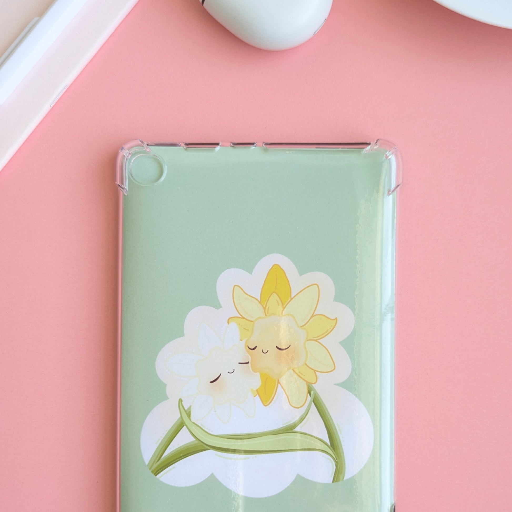 A detailed close-up showing 2/3 of a Kindle with the 'Soft Glow' green kawaii Daffodil Hug Szn insert. The insert is shaped like an oversized die-cut sticker, sitting securely behind a clear case. Soft natural light highlights the high-density gloss, while a pink notepad and white pen are visible in the background, emphasizing a coordinated, monochrome reading ritual.