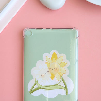 A detailed close-up showing 2/3 of a Kindle with the 'Soft Glow' green kawaii Daffodil Hug Szn insert. The insert is shaped like an oversized die-cut sticker, sitting securely behind a clear case. Soft natural light highlights the high-density gloss, while a pink notepad and white pen are visible in the background, emphasizing a coordinated, monochrome reading ritual.