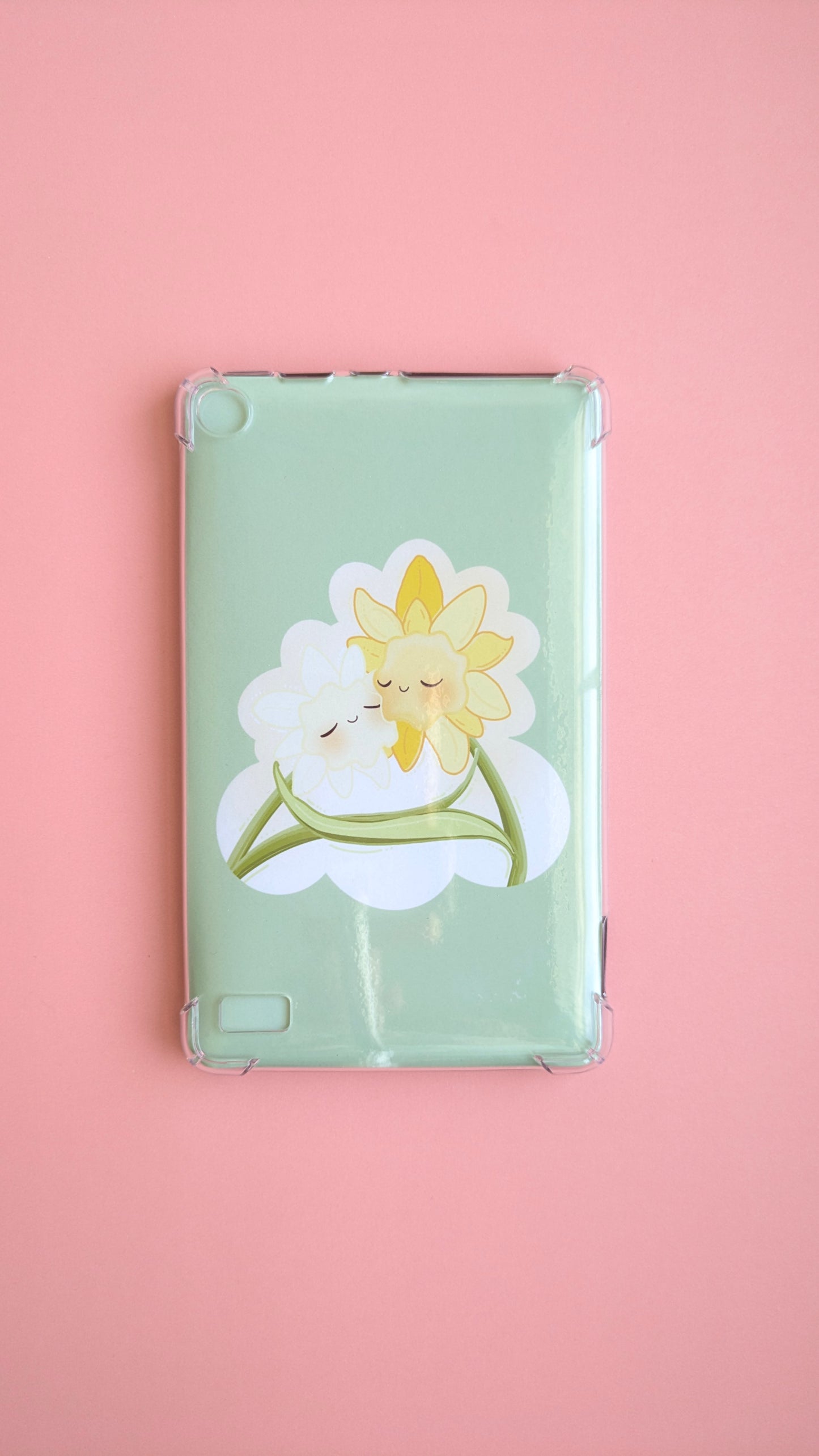 A naturally lit Kindle Paperwhite on a soft pink desk background featuring the 'Soft Glow' green kawaii Daffodil Hug Szn insert. The insert is shaped like an oversized die-cut sticker in warm daffodil yellow, reflecting light off its high-density gloss finish. A clean, adhesive-free tech-scape for intentional reading rituals.