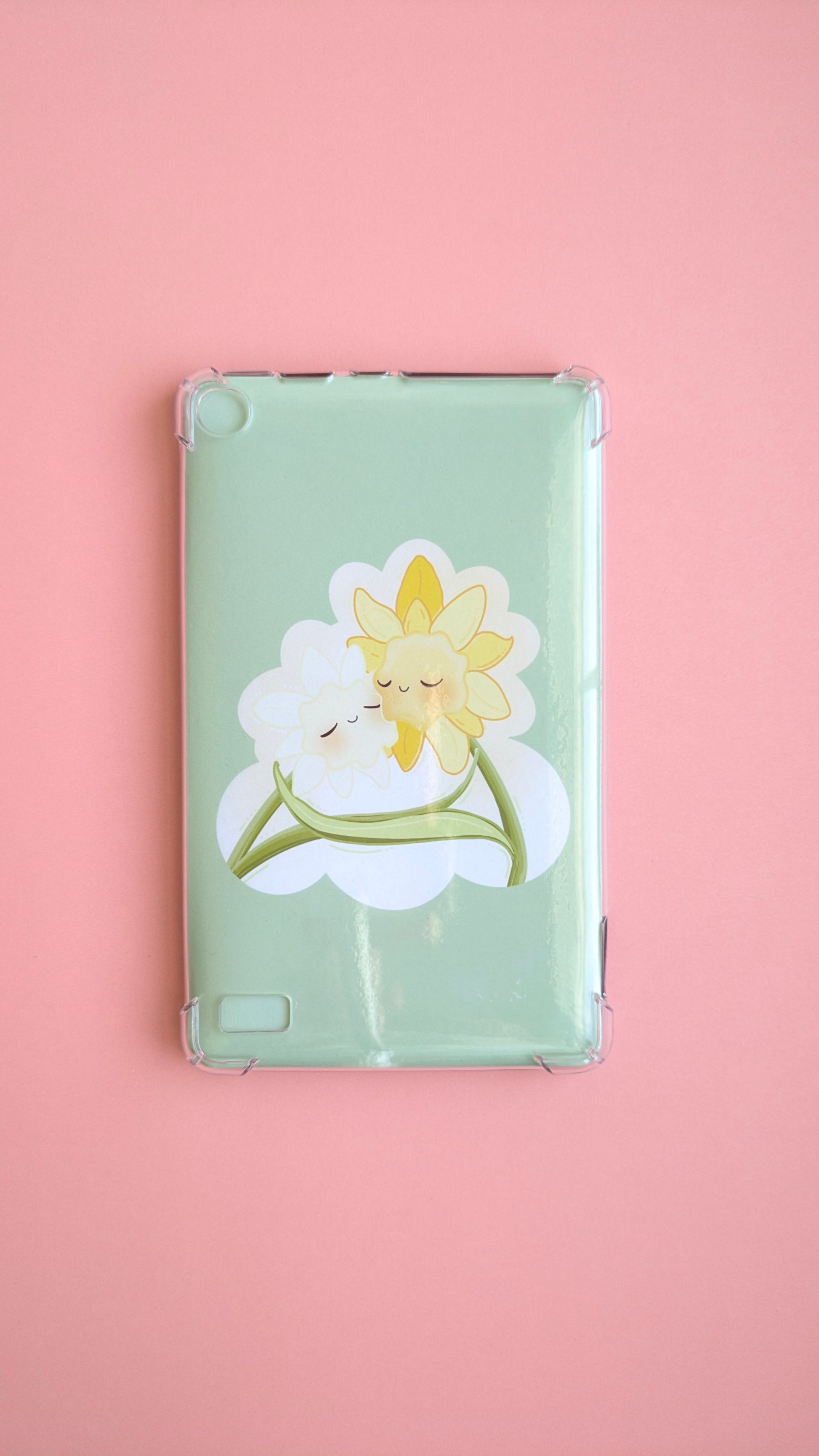 A naturally lit Kindle Paperwhite on a soft pink desk background featuring the 'Soft Glow' green kawaii Daffodil Hug Szn insert. The insert is shaped like an oversized die-cut sticker in warm daffodil yellow, reflecting light off its high-density gloss finish. A clean, adhesive-free tech-scape for intentional reading rituals.