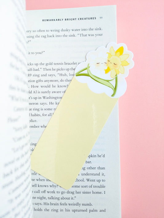 An aerial view of the Soft Glow Yellow Daffodil Hug Szn kawaii bookmark. The die-cut daffodil characters extend over the book edge against a pink background, showcasing a gloss light capture on the laminated finish for a soft pastel reading setup.