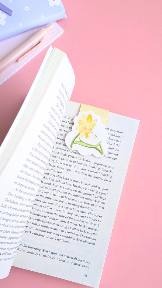 An aerial, angled view of an open copy of 'A Quiet Place' by Seichō Matsumoto, featuring the Daffodil Hug Szn 'Soft Glow' magnetic bookmark. The pale yellow characters are clipped securely to the top of the page, leading the eye down from a pink writing pen and a closed mini notebook in the background. Natural light catches the high-density gloss of the bookmark against the crisp pages, highlighting a coordinated, monochrome-inspired reading ritual for a quiet afternoon.