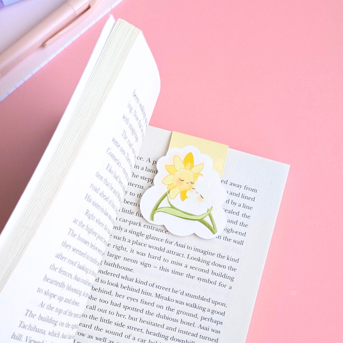 A detailed close-up of the 'Soft Glow' Yellow magnetic bookmark centered on a page of Seichō Matsumoto’s 'A Quiet Place.' The pale yellow kawaii characters stand out against the crisp book text, with a pink writing pen and a lined mini notebook clearly visible in the background. Natural light highlights the high-density gloss finish and secure, page-safe grip, creating a perfectly color-matched stationery aesthetic for intentional journaling and reading.