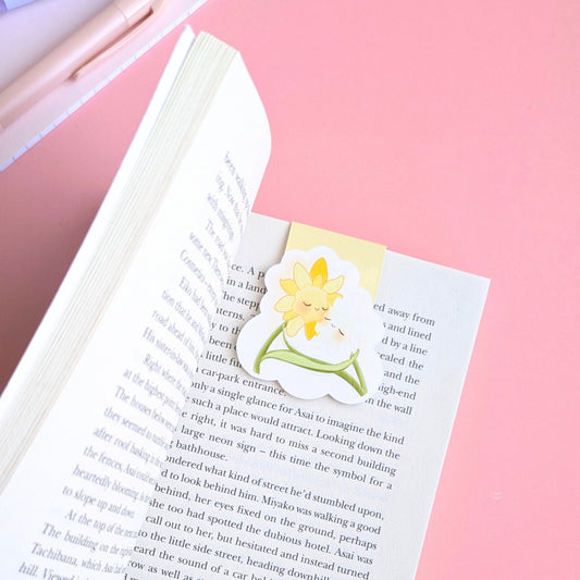 A detailed close-up of the 'Soft Glow' Yellow magnetic bookmark centered on a page of Seichō Matsumoto’s 'A Quiet Place.' The pale yellow kawaii characters stand out against the crisp book text, with a pink writing pen and a lined mini notebook clearly visible in the background. Natural light highlights the high-density gloss finish and secure, page-safe grip, creating a perfectly color-matched stationery aesthetic for intentional journaling and reading.