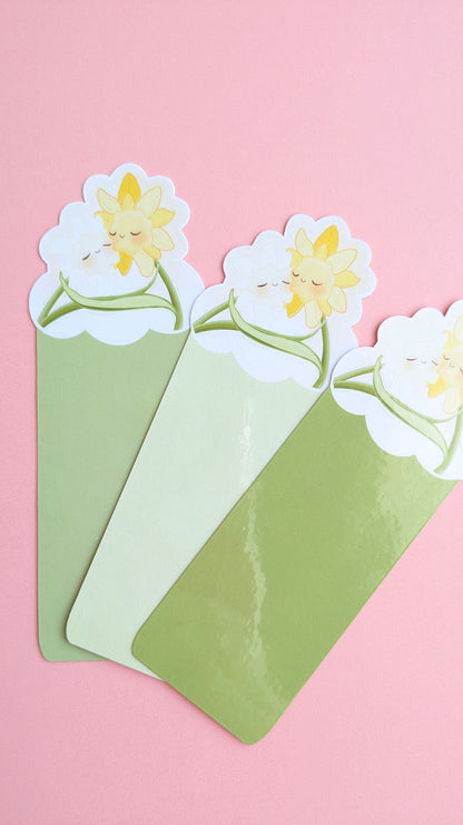 An aerial view of all three green Daffodil Hug Szn bookmarks fanned on a pink background. Vertical Meadow Mist is the base, with Spring Haze as the middle layer, and a high-gloss Meadow Green bookmark on top. Natural light captures a vibrant sheen on the Meadow Green laminate, with a cropped view of the daffodil head on the right.