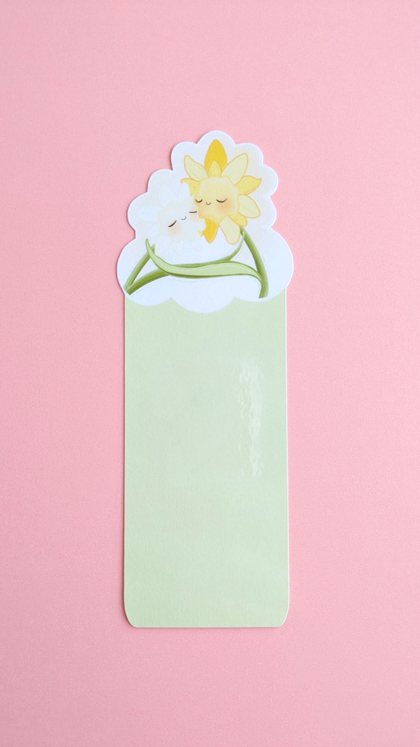 The Daffodil Hug Szn Spring Haze Green daffodil bookmark centered on a pink background. The shot captures a soft gloss sheen on the protective laminate, emphasizing the desaturated, pale green tones of this specific variant for color-obsessed desk-scaping.