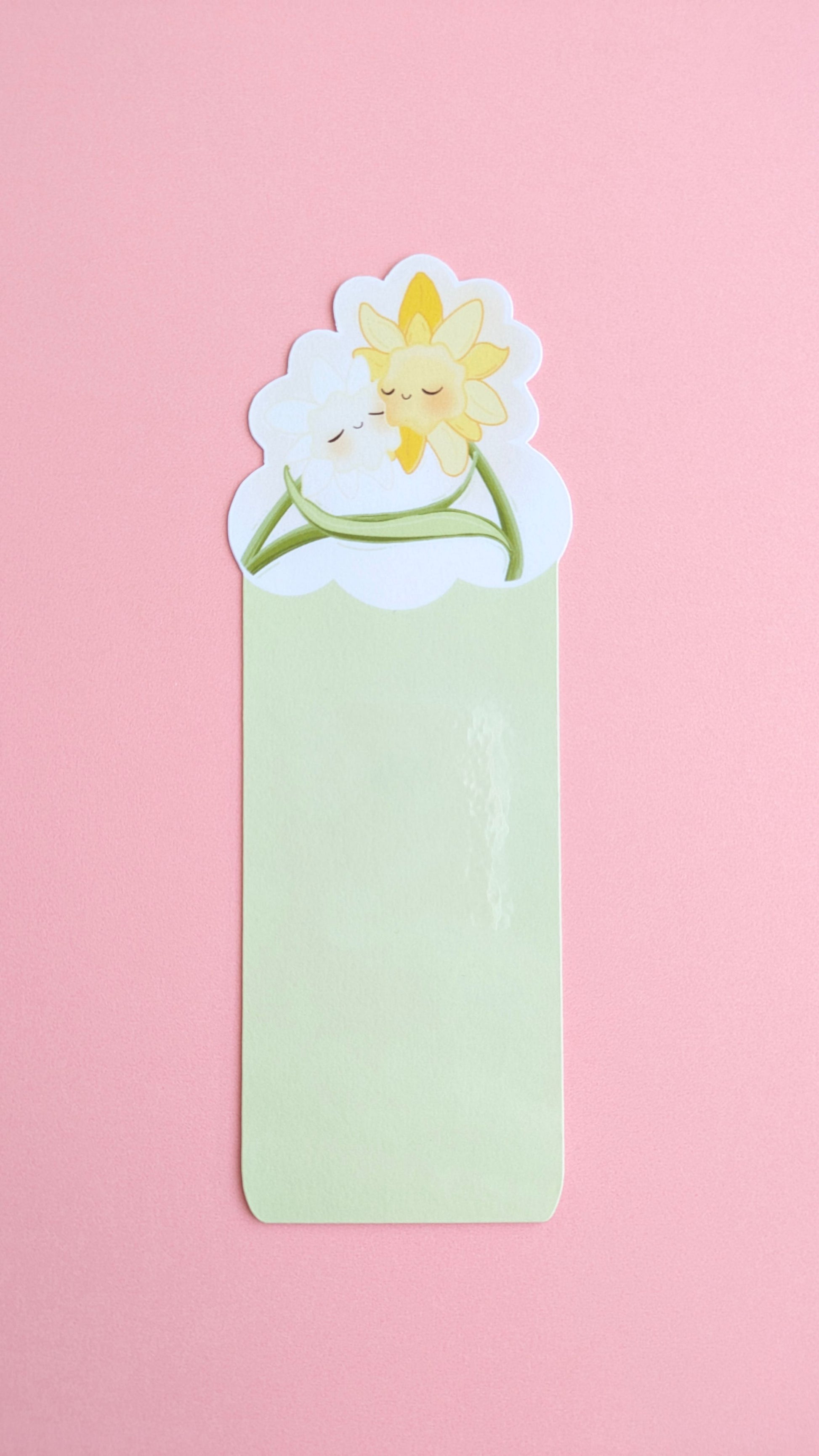 The Daffodil Hug Szn Spring Haze Green daffodil bookmark centered on a pink background. The shot captures a soft gloss sheen on the protective laminate, emphasizing the desaturated, pale green tones of this specific variant for color-obsessed desk-scaping.