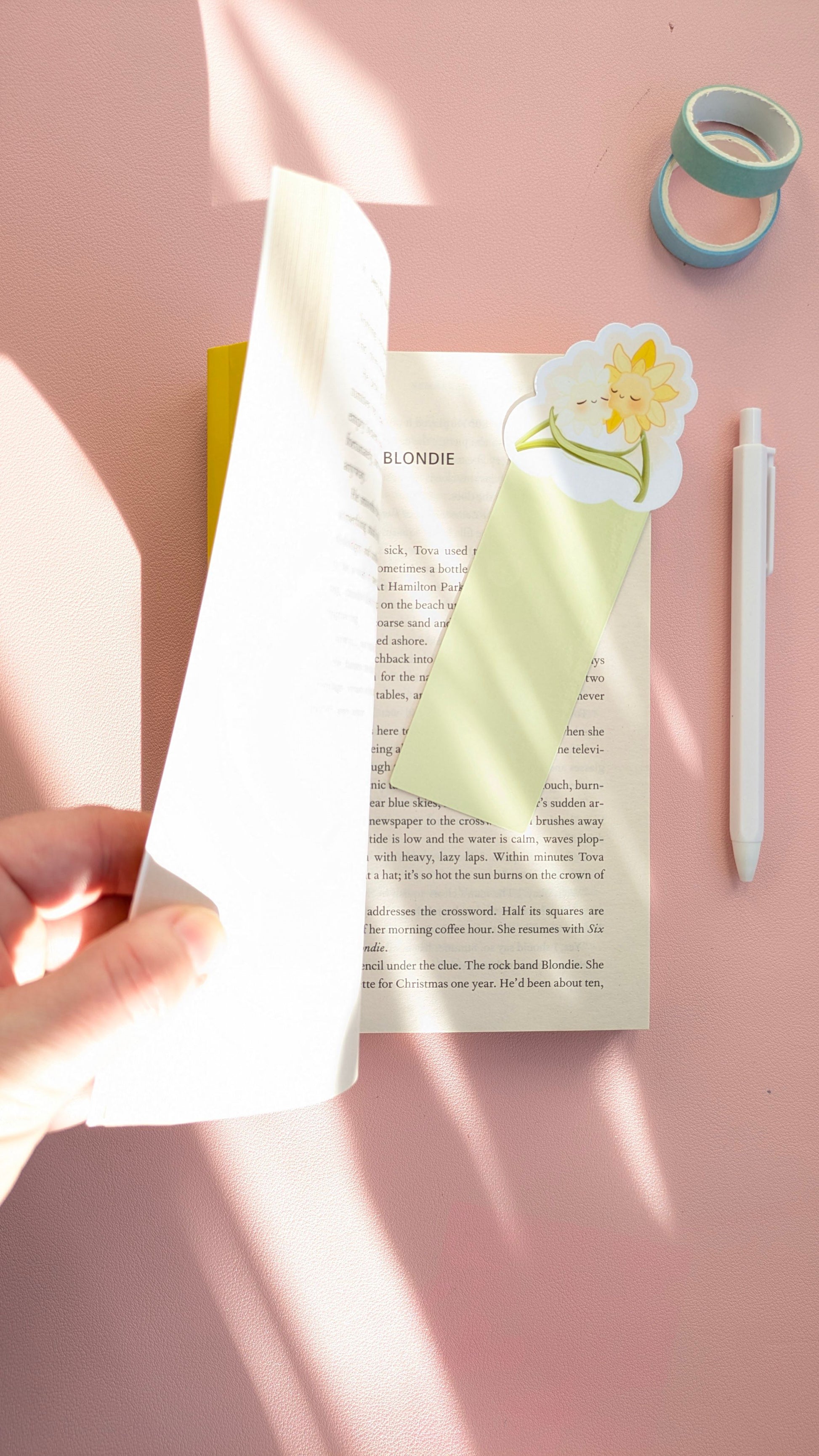 An aerial view of the Daffodil Hug Szn Spring Haze Green bookmark, the lightest green variant in the collection, inside an open book. A hand holds the pages as morning light and window shadows fall across the pink desk, highlighting the pale green die-cut daffodil and an off-white pen.