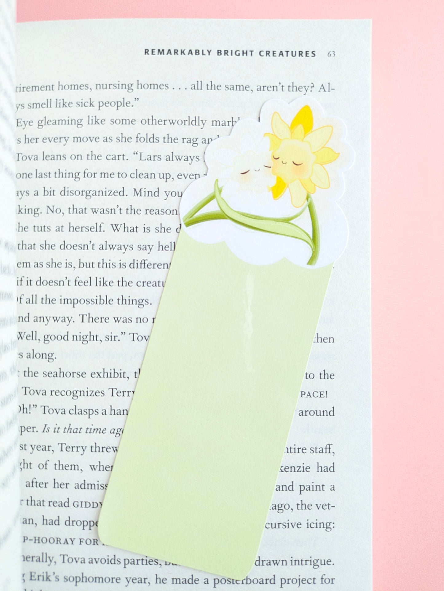 A close-up aerial view of the Daffodil Hug Szn Spring Haze light green daffodil bookmark placed at a slight angle on an open copy of Remarkably Bright Creatures. The composition highlights the soft pastel green tones against a pale pink background and a central gloss sheen on the durable laminate finish.