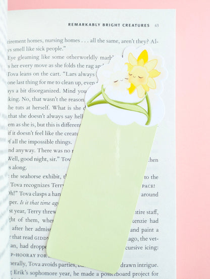 A close-up aerial view of the Daffodil Hug Szn Spring Haze light green daffodil bookmark placed at a slight angle on an open copy of Remarkably Bright Creatures. The composition highlights the soft pastel green tones against a pale pink background and a central gloss sheen on the durable laminate finish.