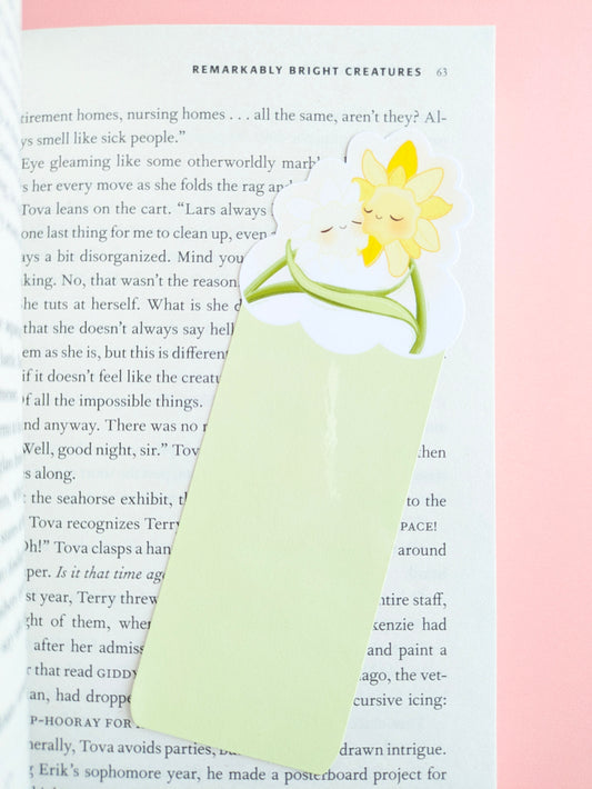 A close-up aerial view of the Daffodil Hug Szn Spring Haze light green daffodil bookmark placed at a slight angle on an open copy of Remarkably Bright Creatures. The composition highlights the soft pastel green tones against a pale pink background and a central gloss sheen on the durable laminate finish.