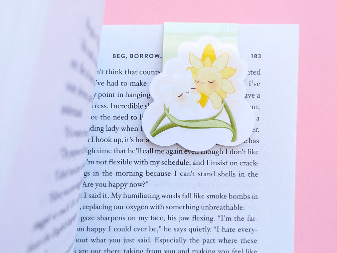 A close-up of a book being opened, revealing a Daffodil Hug Szn 'Spring Haze' magnetic bookmark. The left-hand page of Sarah Adams’ 'Beg, Borrow, or Steal' curls open to reveal the pale, soft-green kawaii character clipped securely to the top right. The high-density gloss finish reflects light off the crisp, curved page, highlighting the secure, no-slide grip for intentional reading rituals.