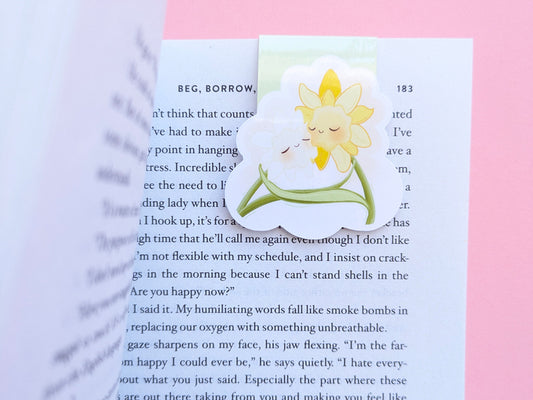 A close-up of a book being opened, revealing a Daffodil Hug Szn 'Spring Haze' magnetic bookmark. The left-hand page of Sarah Adams’ 'Beg, Borrow, or Steal' curls open to reveal the pale, soft-green kawaii character clipped securely to the top right. The high-density gloss finish reflects light off the crisp, curved page, highlighting the secure, no-slide grip for intentional reading rituals.