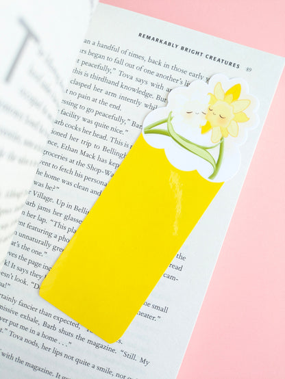 A diagonal overhead view of the Sunny Shine Daffodil Hug Szn kawaii bookmark on a book page. The image features a pink background, a blurred drop cap T on the left page, and a gloss light capture on the vivid yellow base of the die-cut bookmark.