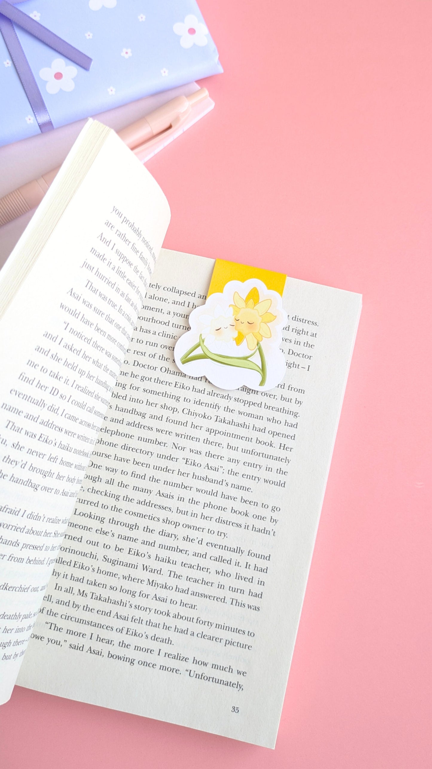 An aerial, naturally lit stationery flat-lay featuring the 'Sunny Shine' yellow magnetic bookmark on an open copy of 'A Quiet Place' by Seichō Matsumoto. In the background, a coordinated gift wrapped in soft blue daisy-print paper with a satin ribbon sits near a pink mini notepad and pen. The rich yellow and kawaii characters anchors the composition, highlighting a color obsessed, ready-to-gift aesthetic for intentional readers and stationery collectors.