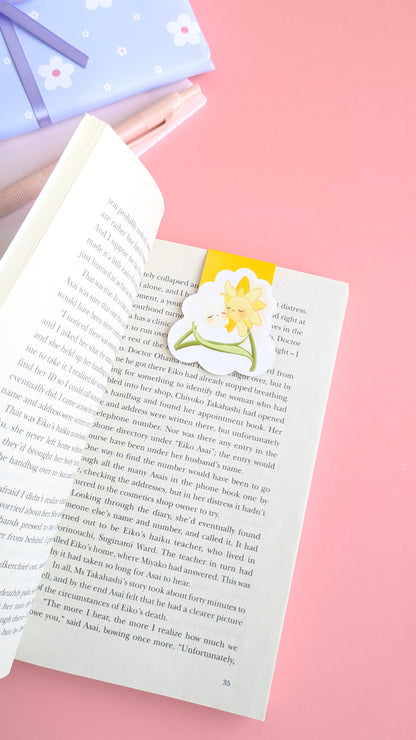 An aerial, naturally lit stationery flat-lay featuring the 'Sunny Shine' yellow magnetic bookmark on an open copy of 'A Quiet Place' by Seichō Matsumoto. In the background, a coordinated gift wrapped in soft blue daisy-print paper with a satin ribbon sits near a pink mini notepad and pen. The rich yellow and kawaii characters anchors the composition, highlighting a color obsessed, ready-to-gift aesthetic for intentional readers and stationery collectors.