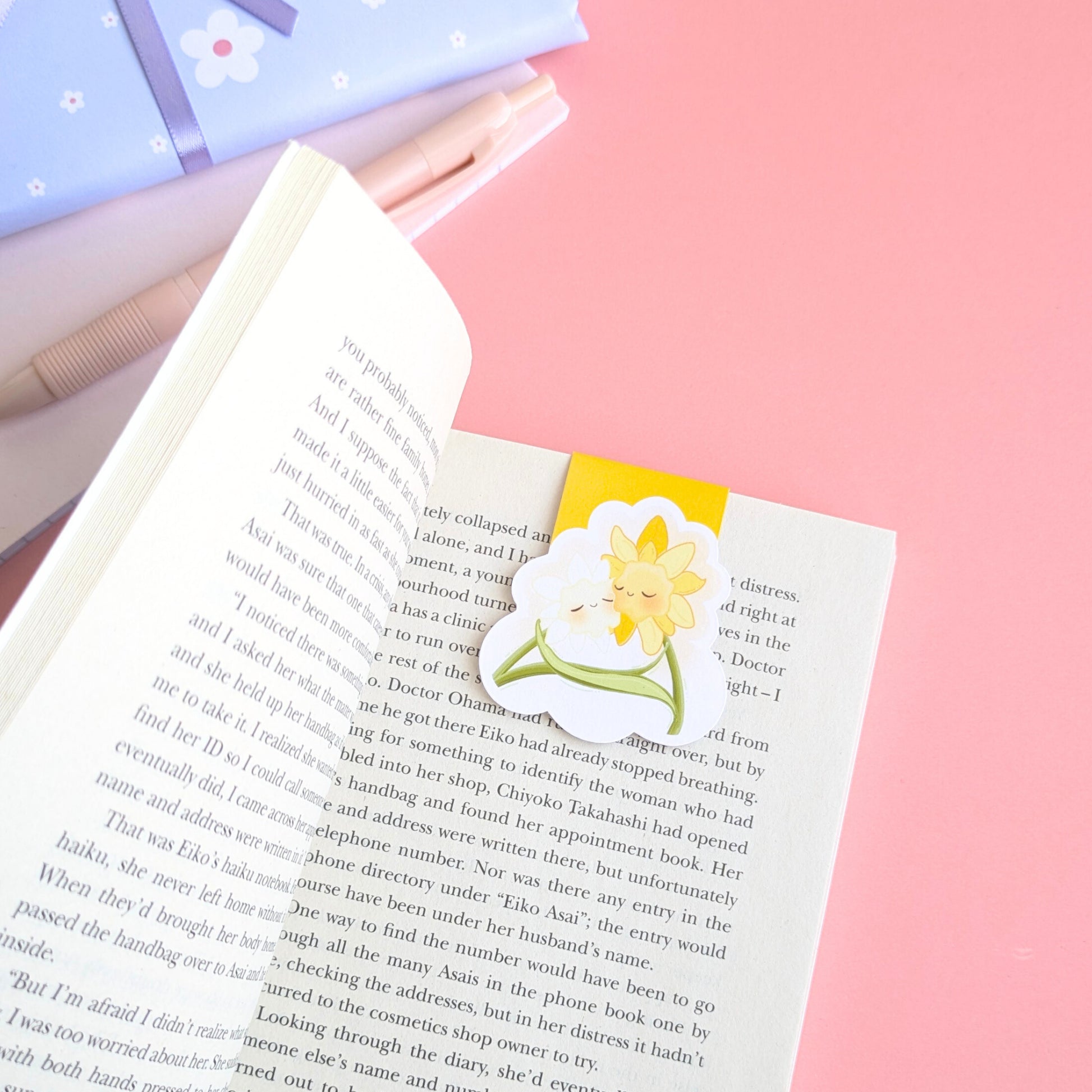 A close-up, high-angle view of the 'Sunny Shine' yellow Daffodil Hug Szn magnetic bookmark centered on a curved book page. The vibrant yellow reflects natural light off its gloss finish, showing a secure, page safe grip. A pink notepad and a hint of blue daisy patterned gift wrap are visible in the soft-focus background, emphasizing the premium material quality and intentional color matching of the Daffodil Hug Szn collection.