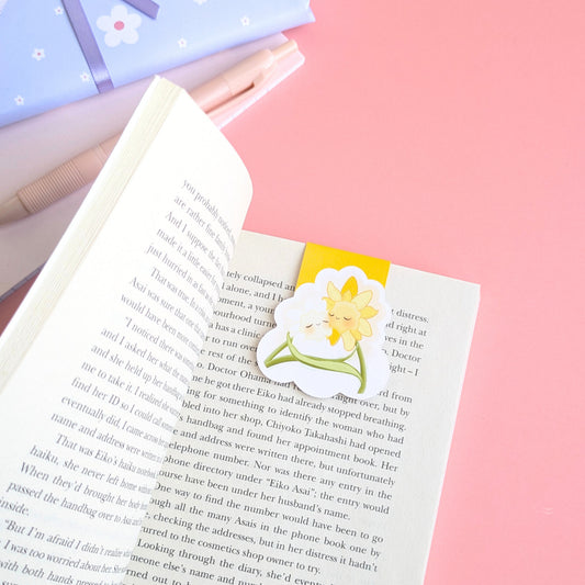 A close-up, high-angle view of the 'Sunny Shine' yellow Daffodil Hug Szn magnetic bookmark centered on a curved book page. The vibrant yellow reflects natural light off its gloss finish, showing a secure, page safe grip. A pink notepad and a hint of blue daisy patterned gift wrap are visible in the soft-focus background, emphasizing the premium material quality and intentional color matching of the Daffodil Hug Szn collection.