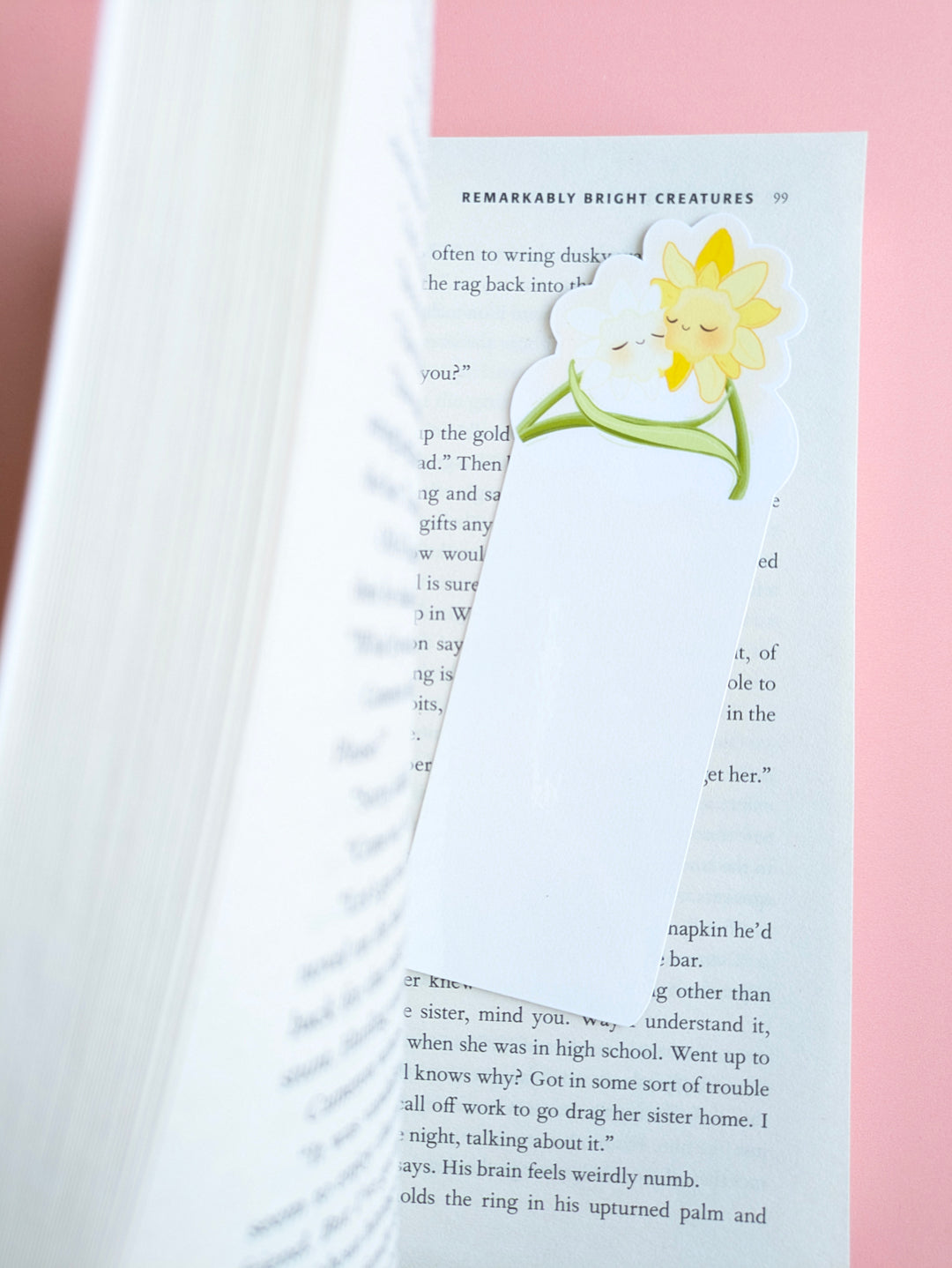 An aerial view of the kawaii White Bloom Daffodil Hug Szn bookmark placed inside a book opened two-thirds of the way. The white die-cut daffodils are angled on a pink background, with thick pages on the left creating a curved shadow and a gloss light tint visible on the laminated surface.