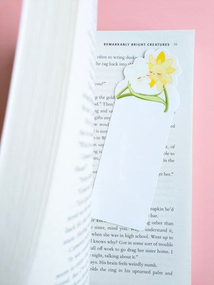 An aerial view of the kawaii White Bloom Daffodil Hug Szn bookmark placed inside a book opened two-thirds of the way. The white die-cut daffodils are angled on a pink background, with thick pages on the left creating a curved shadow and a gloss light tint visible on the laminated surface.