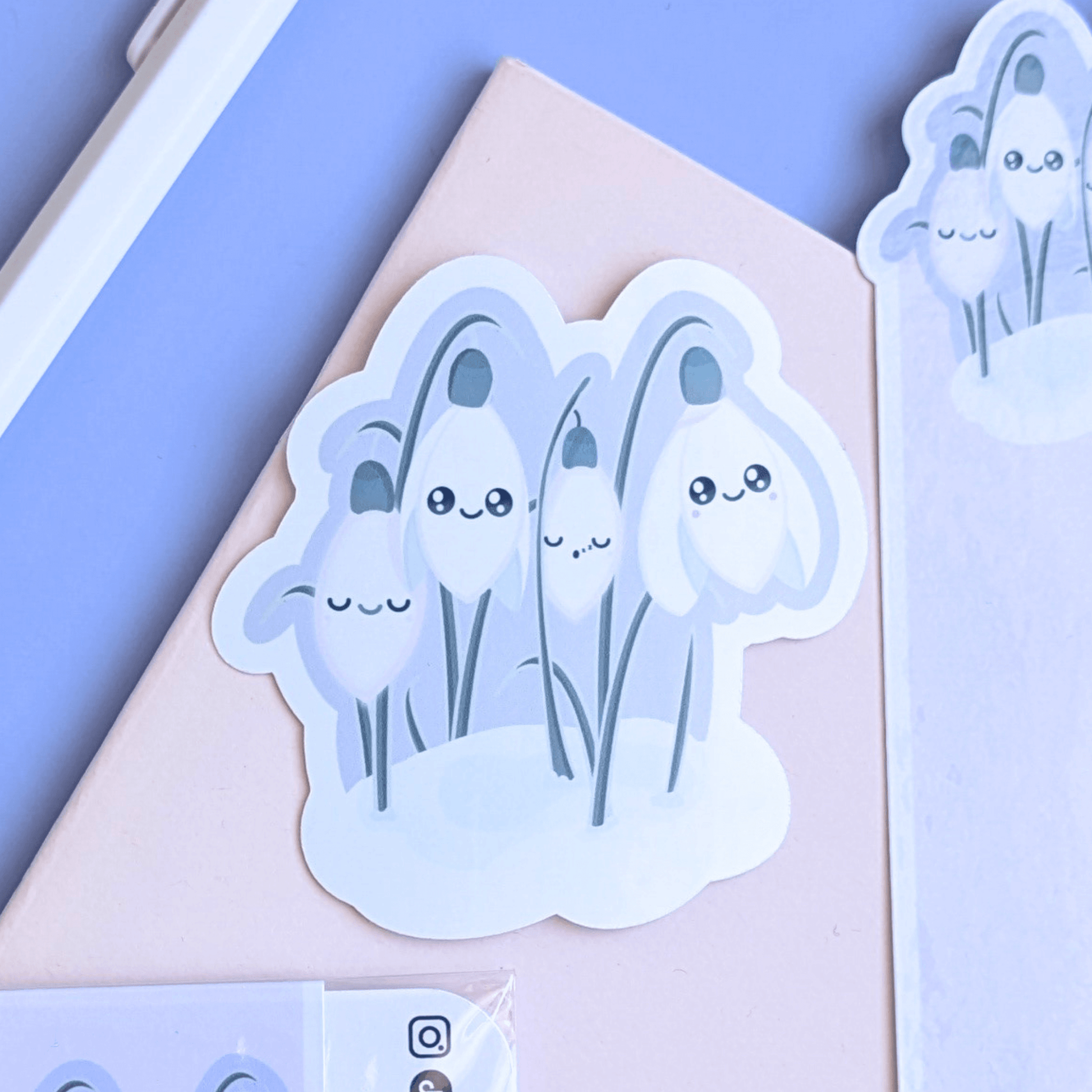 Detailed close-up of a 3-inch Sleepy Snowdrop 'Dawn' Kawaii sticker on a pink pocket notebook. The character art features Snowdrop White blooms with sage-silver leaves and a cozy soft-glow effect against a lavender-blue dawn sky. This gloss decal is the focal point of a mindful journaling setup, showcasing the quintessential Kawaii faces and color-coordinated palette for a visual life-building aesthetic.