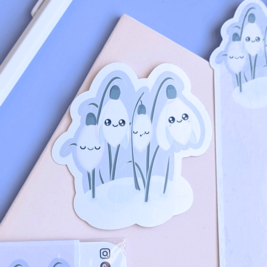 Detailed close-up of a 3-inch Sleepy Snowdrop 'Dawn' Kawaii sticker on a pink pocket notebook. The character art features Snowdrop White blooms with sage-silver leaves and a cozy soft-glow effect against a lavender-blue dawn sky. This gloss decal is the focal point of a mindful journaling setup, showcasing the quintessential Kawaii faces and color-coordinated palette for a visual life-building aesthetic.