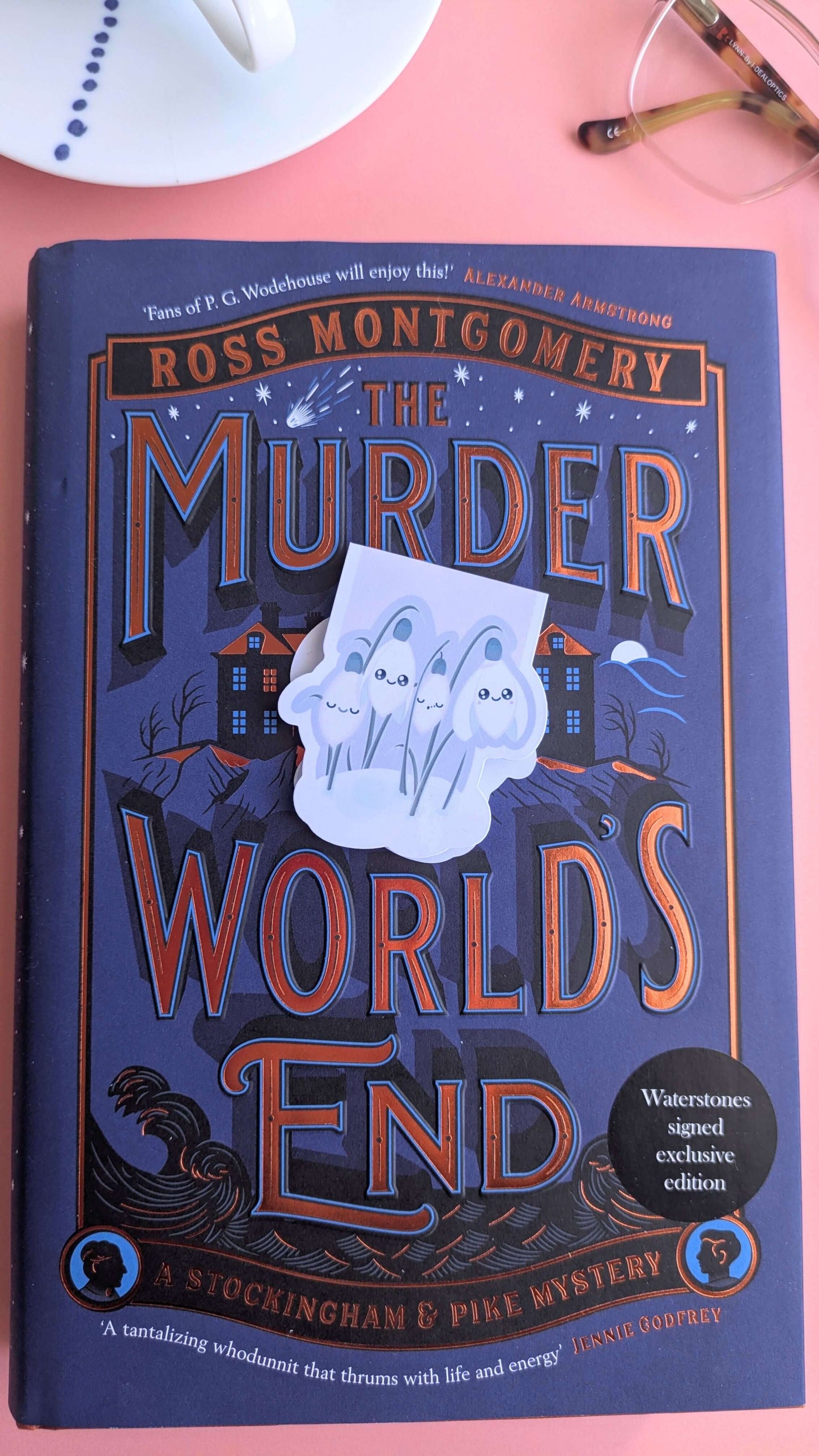 The Kawaii Sleepy Snowdrop 'Dawn' lavender blue magnetic bookmark angled to the right in the center of a signed exclusive edition book cover. Styled with a French porcelain cup and saucer showing hand-painted blue dot details, highlighting a mindful and aesthetic morning reading ritual.