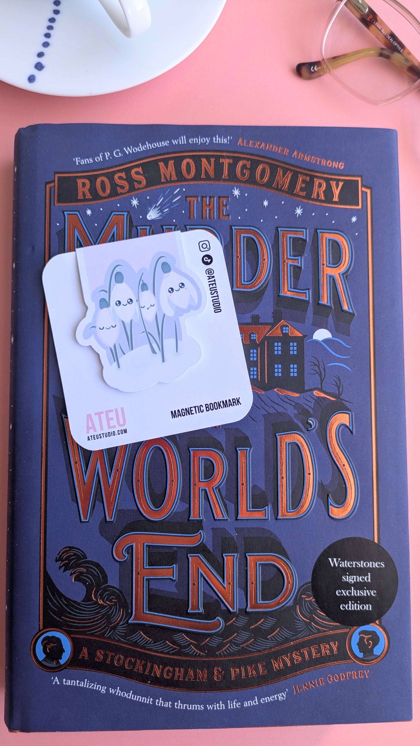 An Ateu Studio Kawaii Sleepy Snowdrop 'Dawn' lavender blue magnetic bookmark on its backing card, offset at a right angle over the cover of 'The Murder at World’s End' by Ross Montgomery. This Waterstones signed exclusive edition with sprayed gold edges is styled on a pink background with a white French porcelain cup and reading glasses.