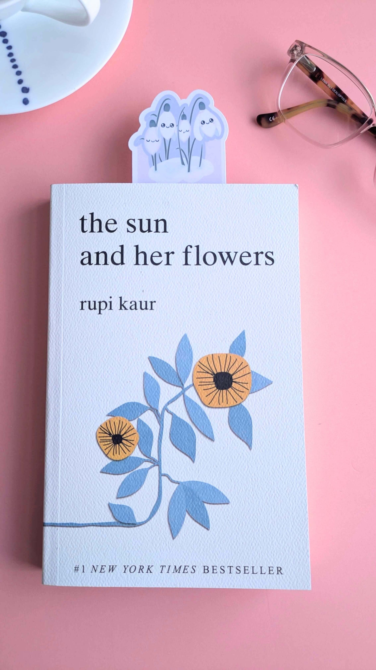 An Ateu Studio Kawaii Sleepy Snowdrop 'Dawn' lavender blue bookmark protruding from the top of a closed copy of Rupi Kaur’s 'The Sun and Her Flowers'. The die-cut snowdrop cluster and little snow pile are clearly visible against the book cover and soft rose-pink background. Styled with a white porcelain cup and reading glasses under morning window light, emphasizing a peaceful, aesthetic reading ritual.