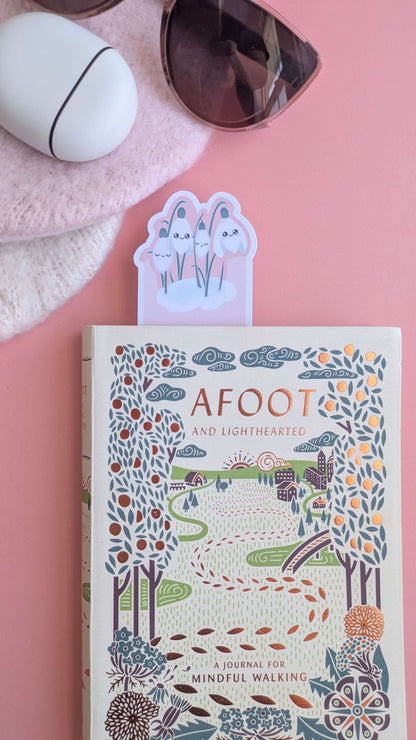 A Kawaii Sleepy Snowdrop 'Icy Venusian Pink' bookmark peeking from a closed copy of 'Afoot and Lighthearted: A Journal for Mindful Walking'.  This intentional, color-coordinated scene styled with Fossil-style translucent sunglasses and a soft pink hat, on a rose-pink background highlights the unique die-cut shape and Kawaii character art for aesthetic book decorating and visual life building.