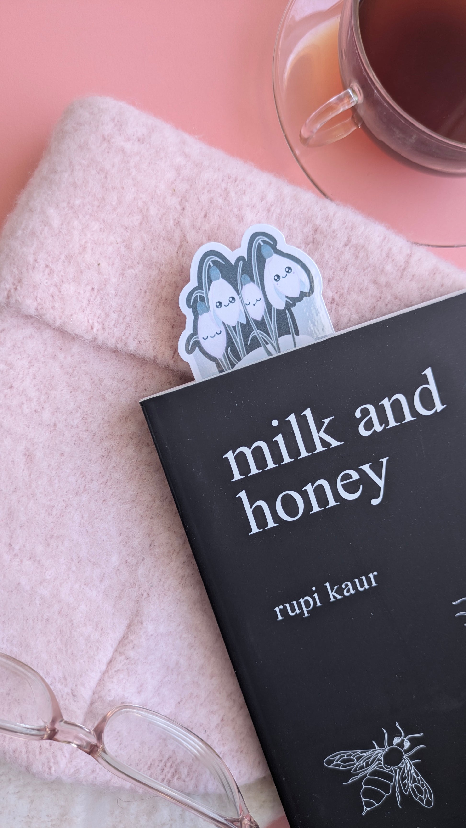 A Kawaii Sleepy Snowdrop 'Winter’s Edge' teal bookmark protruding from the top of a closed copy of Rupi Kaur’s 'Milk and Honey'. The die-cut snowdrop cluster and snowy patch are clearly visible against the black book cover and soft pink wool hat. The composition features the reading glasses and glass French champagne-region cup and saucer, emphasizing a cohesive, cool-toned winter aesthetic.