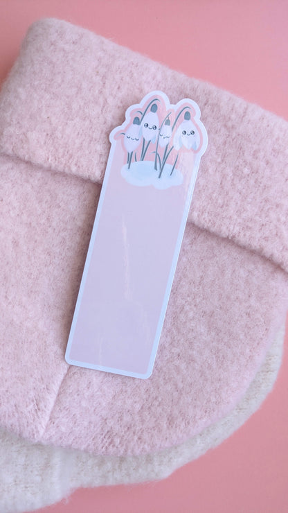 A Kawaii Sleepy Snowdrop 'Icy Venusian Pink' die-cut bookmark centered on an elegant soft pink wooly winter hat. The shot captures multiple reflective gloss sheen points across the laminated surface, highlighting the Snowdrop White characters and sage-silver leaves. A hint of an off-white scarf underneath completes this quintessentially kawaii, cozy, romanticized winter reading accessory scene.