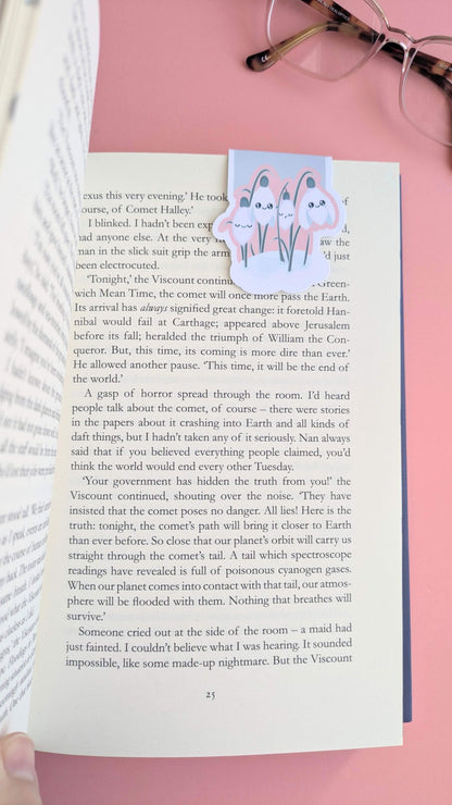 An aerial shot of an Ateu Studio Kawaii Sleepy Snowdrop 'Icy Venusian' pink magnetic bookmark on the right-hand page of a freshly opened book. A thumb is visible at the spine, capturing the moment of opening a new story, with the pages showing a natural lift at the bottom. The scene is styled with reading glasses on a rose-pink background in natural light, emphasizing the soft pink tones and the tactile, intentional experience of a new reading journey.